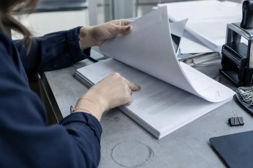 Hands turning to the last page of a stapled signed contract on a cluttered desk