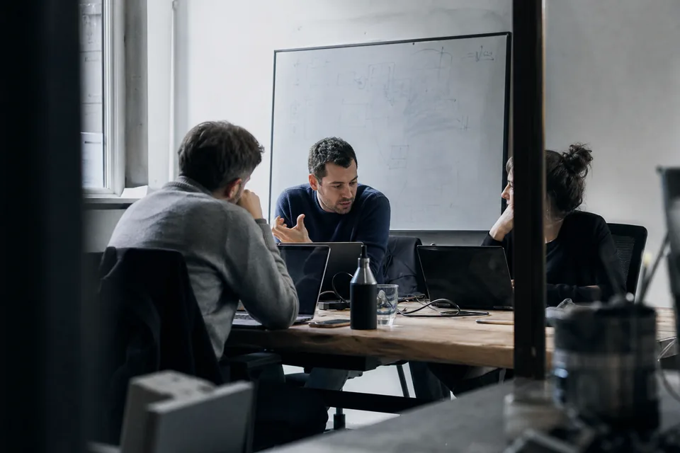Three colleagues discussing work at a wooden table with laptops
