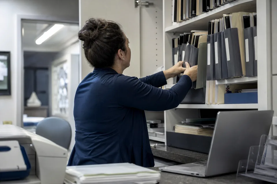 Office manager reorganising document folders on a shelf next to a laptop